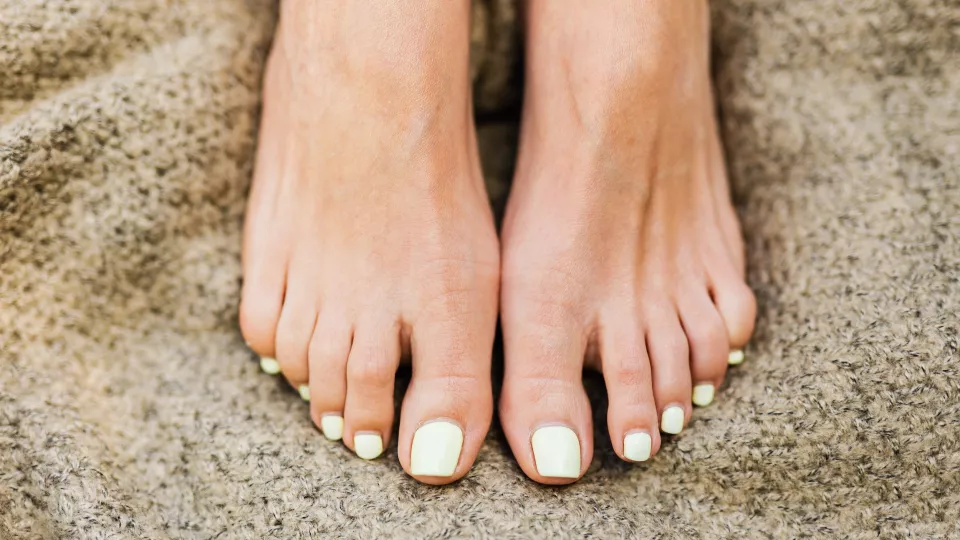 Fötter med gult nagellack vilar på en sandfärgad filt. Foto.
