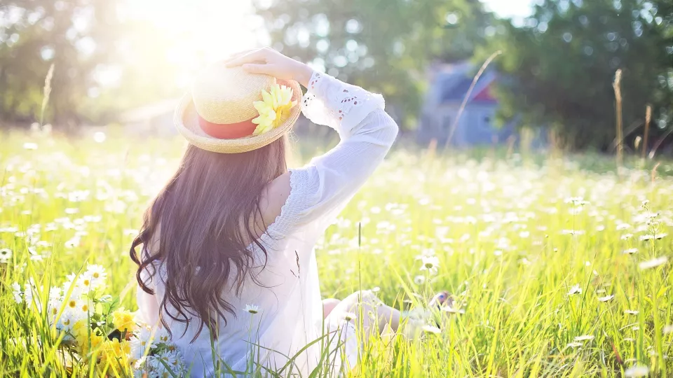Foto på en kvinna sedd bakifrån med brunt hår och en gul hatt som som sitter ner i gräs och tittar mot solen som skiner starkt.