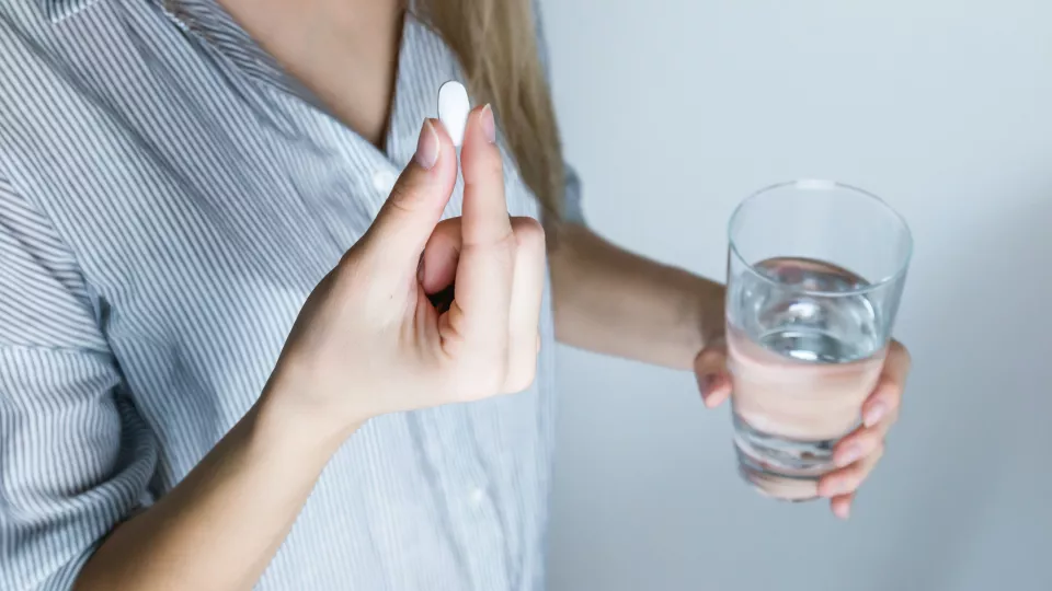 Närbild på person som som håller i en vit tablett i ena handen och ett glas vatten i den andra. 
