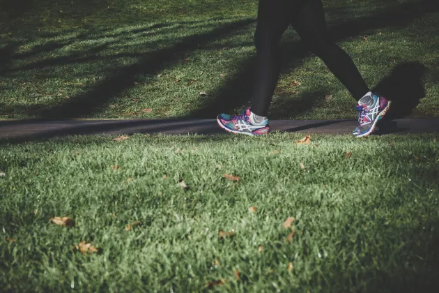 En person i svarta träningsbyxor och flerfärgade gympaskor som går längs en gångväg som ligger mellan två gräsmattor. Endast personens underkropp syns.