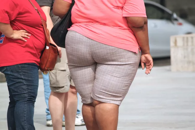 En överviktig kvinna i jeans och röd t-shirt står med händerna på midjan. Bredvid henne går en överviktig kvinna i shorts och rosa t-shirt.