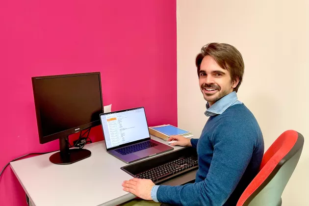 Ett foto på Simone som sitter på en röd datorstol vid sitt skrivbord framför sin laptop. Skrivbordet står mot en rosa vägg. Simone har brunt hår och har en blå skjorta på sig under en blå pullover. Han ler.