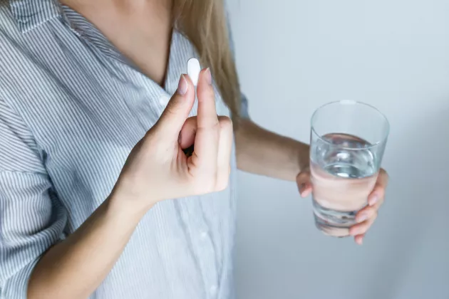Närbild på person som som håller i en vit tablett i ena handen och ett glas vatten i den andra. 