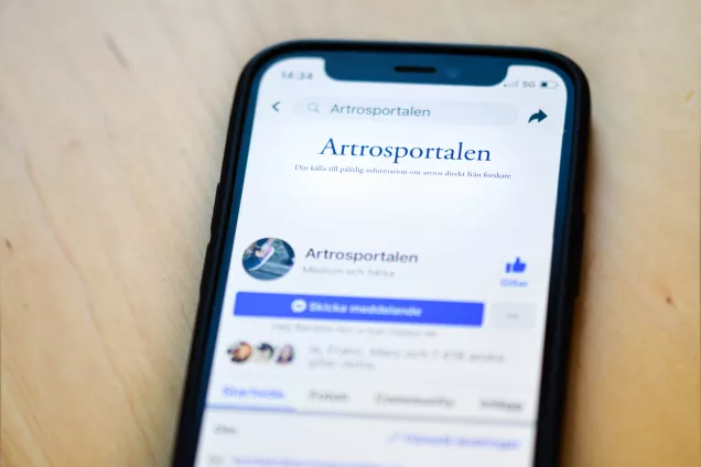 Foto på en smartphone med Artrosportalens Facebook-sida som bakgrund. Mobiltelefonen ligger mot en träbakgrund.