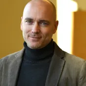 Porträttbild på Martin Englund. Martin är en vit man och har på sig en grå polotröja och en grön kavaj.