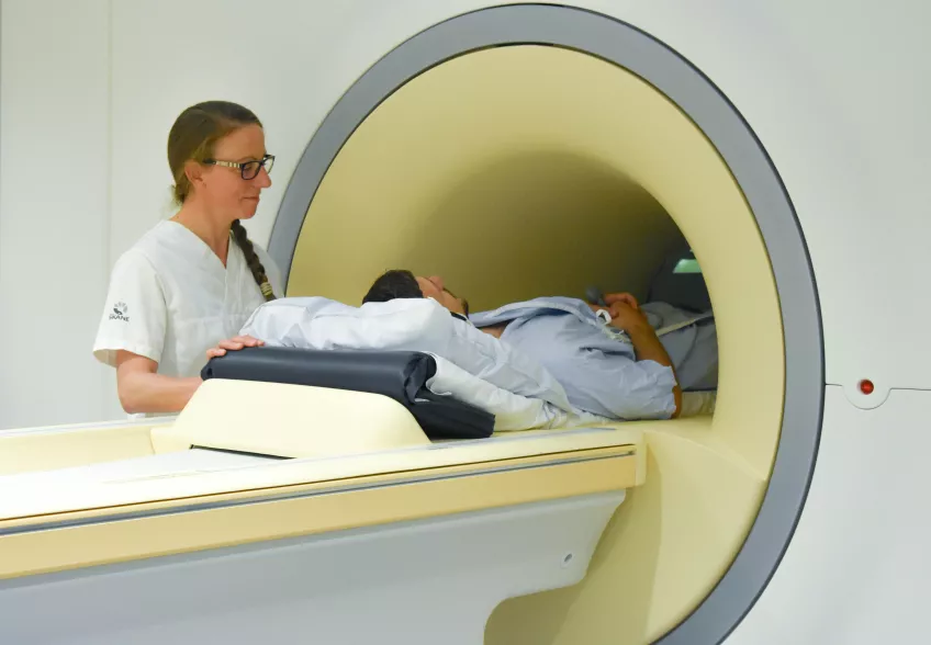 En kvinna i vita sjukhuskläder står bredvid en man som ligger ner med benen i en magnetröntgen.