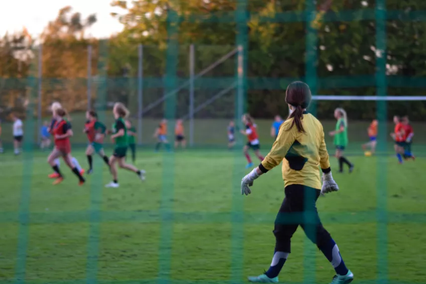 En actionbild på en kvinnlig fotbollsmålvakt i gul tröja och svarta byxor som går och tittar ut mot ett gäng kvinnliga fotbollsspelare på fotbollsplanen. Fotografiet är taget genom fotbollsmålsnätet.