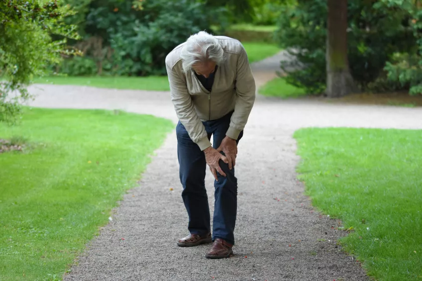 En äldre man med grått hår, blåa byxor och beige jacka står upp med lätt böjning och håller i sitt knä. Mannen står på en grusväg med gräs på vardera sida av vägen.