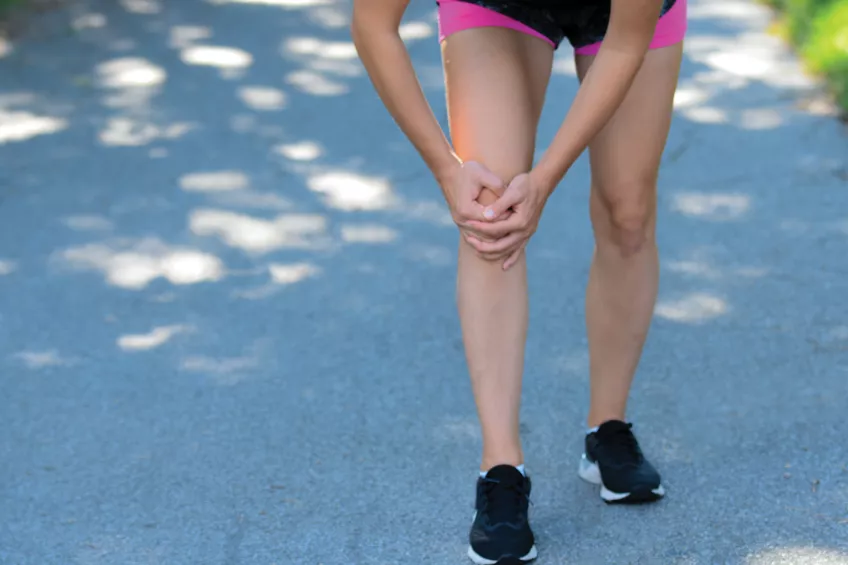 En ung kvinna i rosa och svarta shorts och svarta träningsskor som håller i sitt ena knä. Endast underkroppen syns.