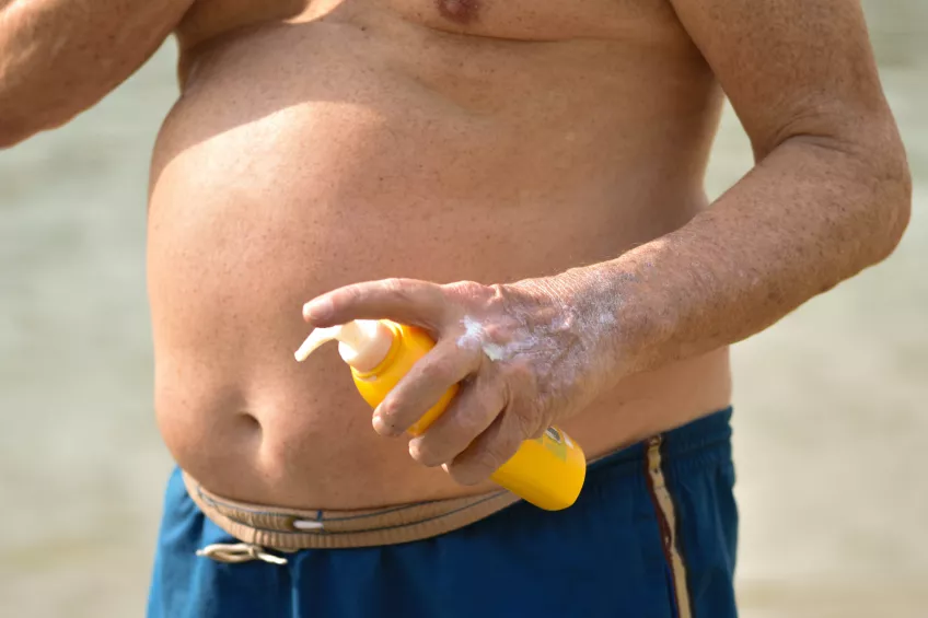 Magen på en överviktig vit man i blåa badshorts. Mannen håller i en gul flaska med solkräm.