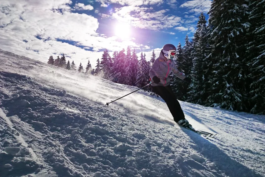 En kvinna i skidkläder, skidhjälm och skidglasögon som åker skidor ner för en snöig backe.