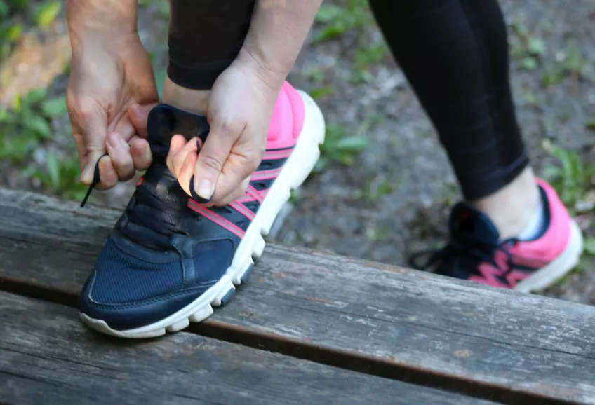 En inzoomad bild på en persons händer som knyter sin blå-rosa träningssko. Benet med skon som knyts är upphöjt på en träbänk medan det andra benet är kvar på marken. Foto.