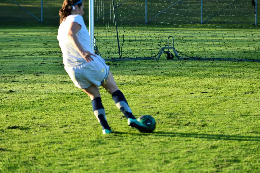 En actionbild på en kvinna i vita fotbollskläder som sparkar en fotboll in i ett fotbollsmål på en gräsplan