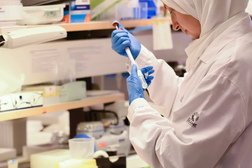 En kvinna i en vit labbrock med blåa plasthandskar håller i en pipett och ett provrör inuti ett laboratorium.
