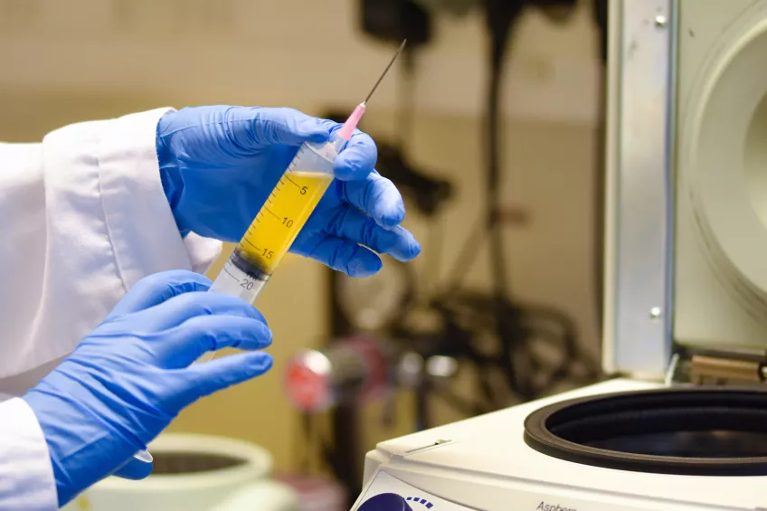 Ett par händer i blåa plasthandskar håller i en spruta fylld med gul blodplasma framför en centrifug.