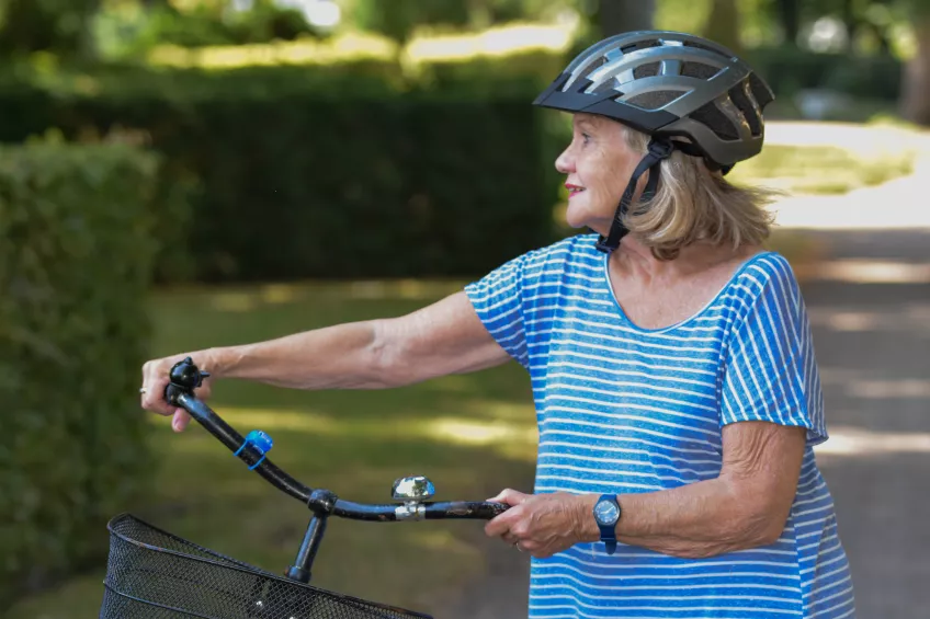 En kvinna i blå- och vitrandig t-shirt och silvrig cykelhjälm står och håller i en cykel och tittar till sidan. Foto.