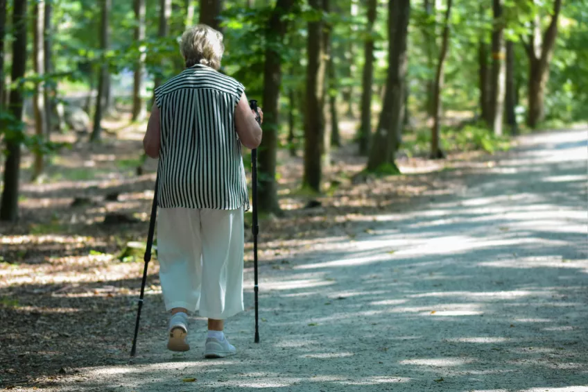 Foto på en kvinna i vita byxor och randig topp som går stavgång längs en grusväg i skogen.