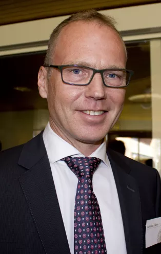 Porträttbild på Carl Johan Tiderius. Carl Johan är en vit man med glasögon som har på sig en vit skjorta, svart kavaj och en mörkblå slips med röda prickar.