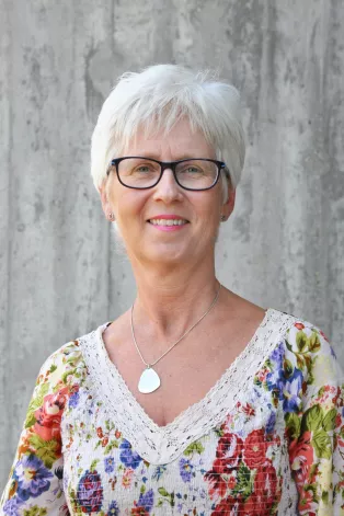Porträtt på Eva Ekvall Hansson. Eva är en vit kvinna med kort hår och glasögon som har på sig en blommig blus.